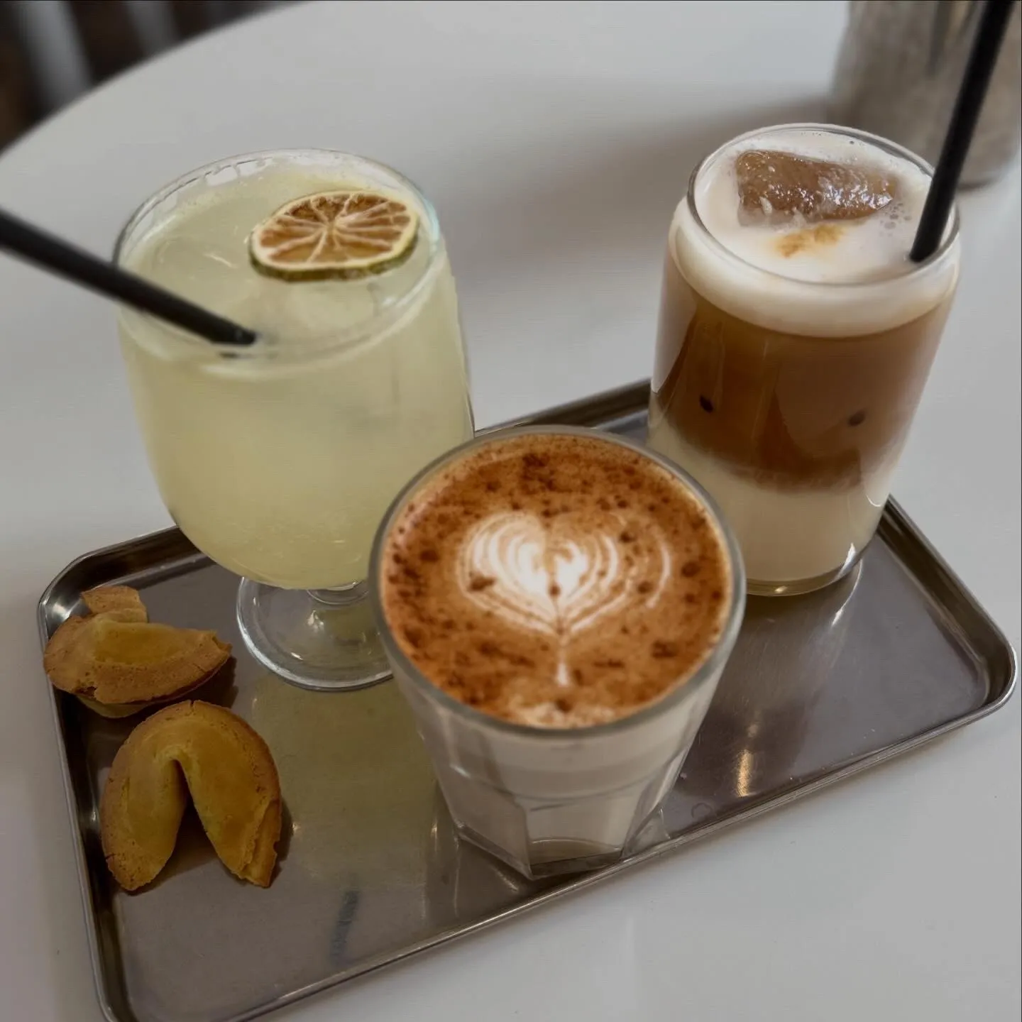 Un café con leche, un café frío y una limonada en una bandeja de metal, con galletas de la fortuna